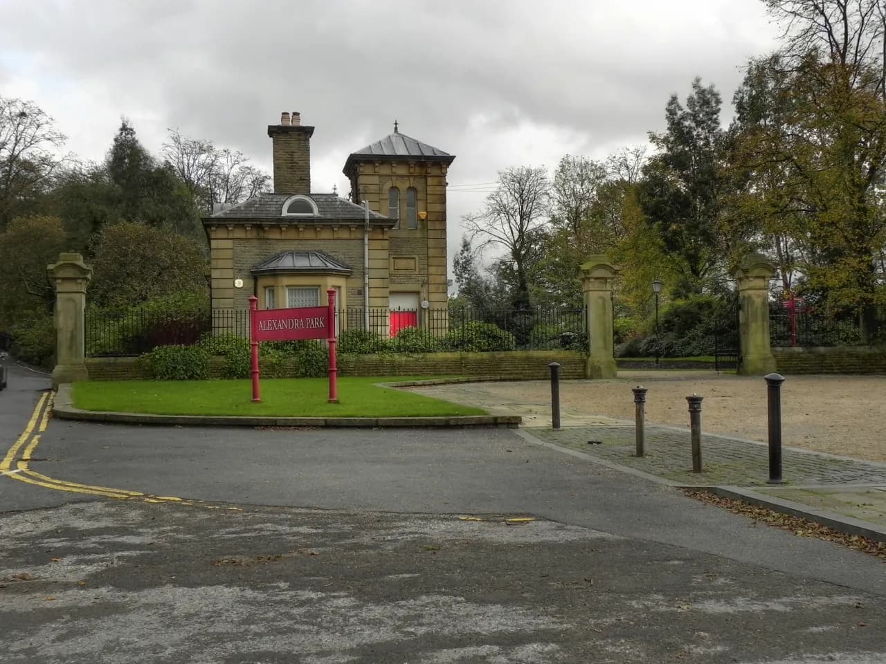 Entrance to Alexandra Park with a stone building, a red sign, and a paved area under an overcast sky.