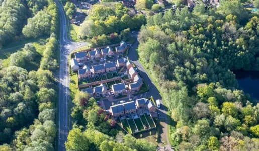 An aerial view of a housing development surrounded by lush green trees and a road, with a body of water visible in the upper right corner.