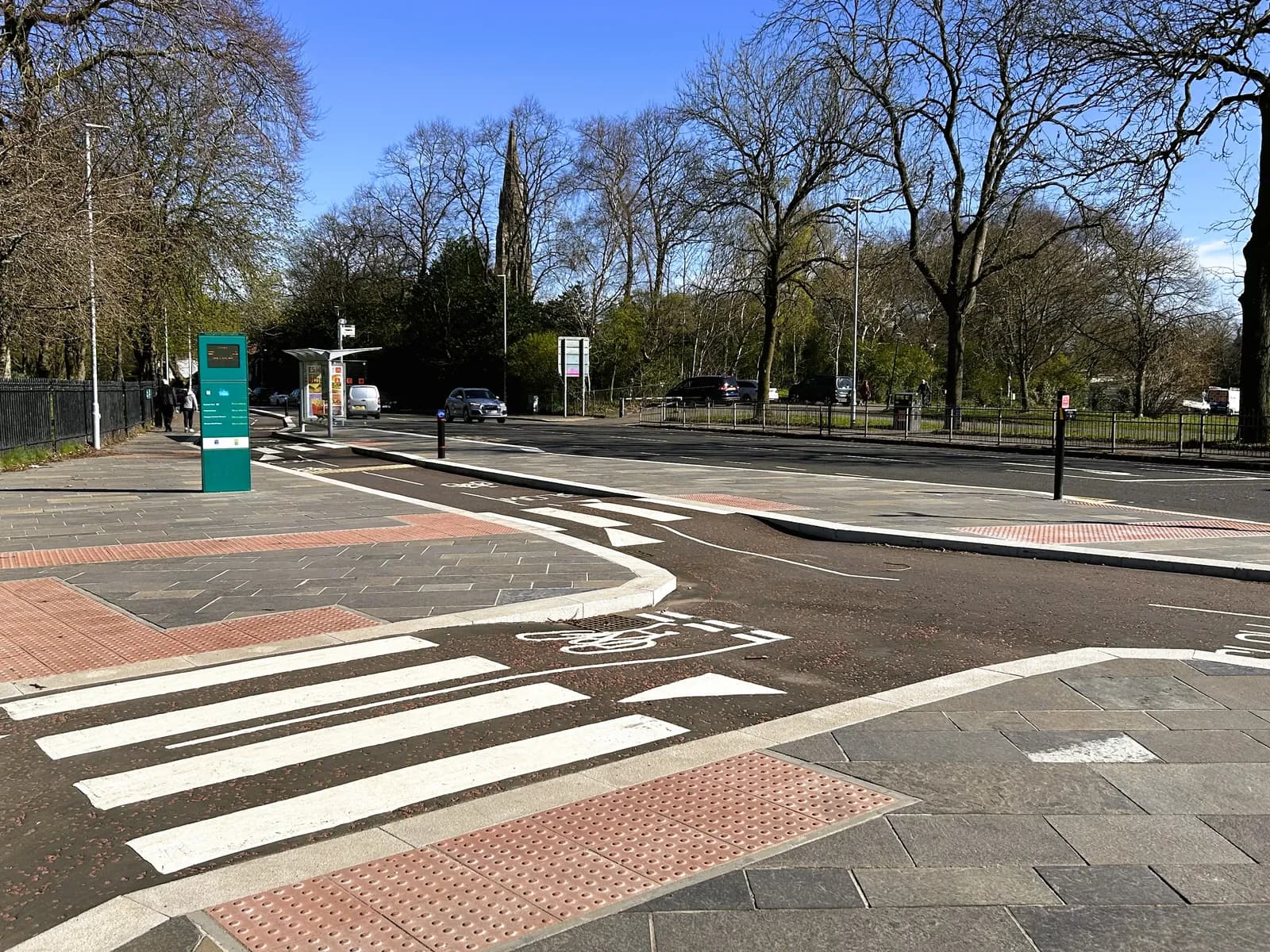 A newly installed pedestrian and cycle crossing with clear white markings, red tactile paving, and a green information display box on the left, next to a tree-lined street.