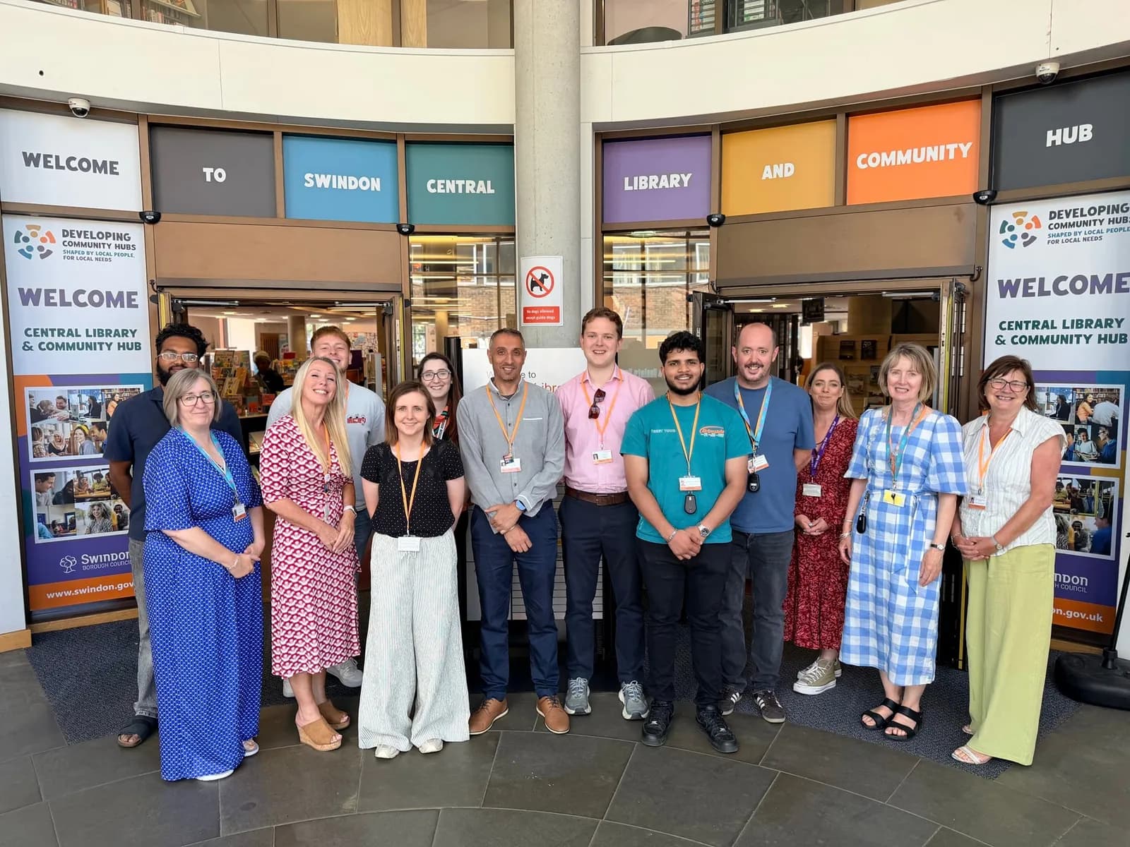 A group of diverse people stand smiling in front of a library entrance with signs above them that read 'WELCOME TO SWINDON CENTRAL LIBRARY AND COMMUNITY HUB'.