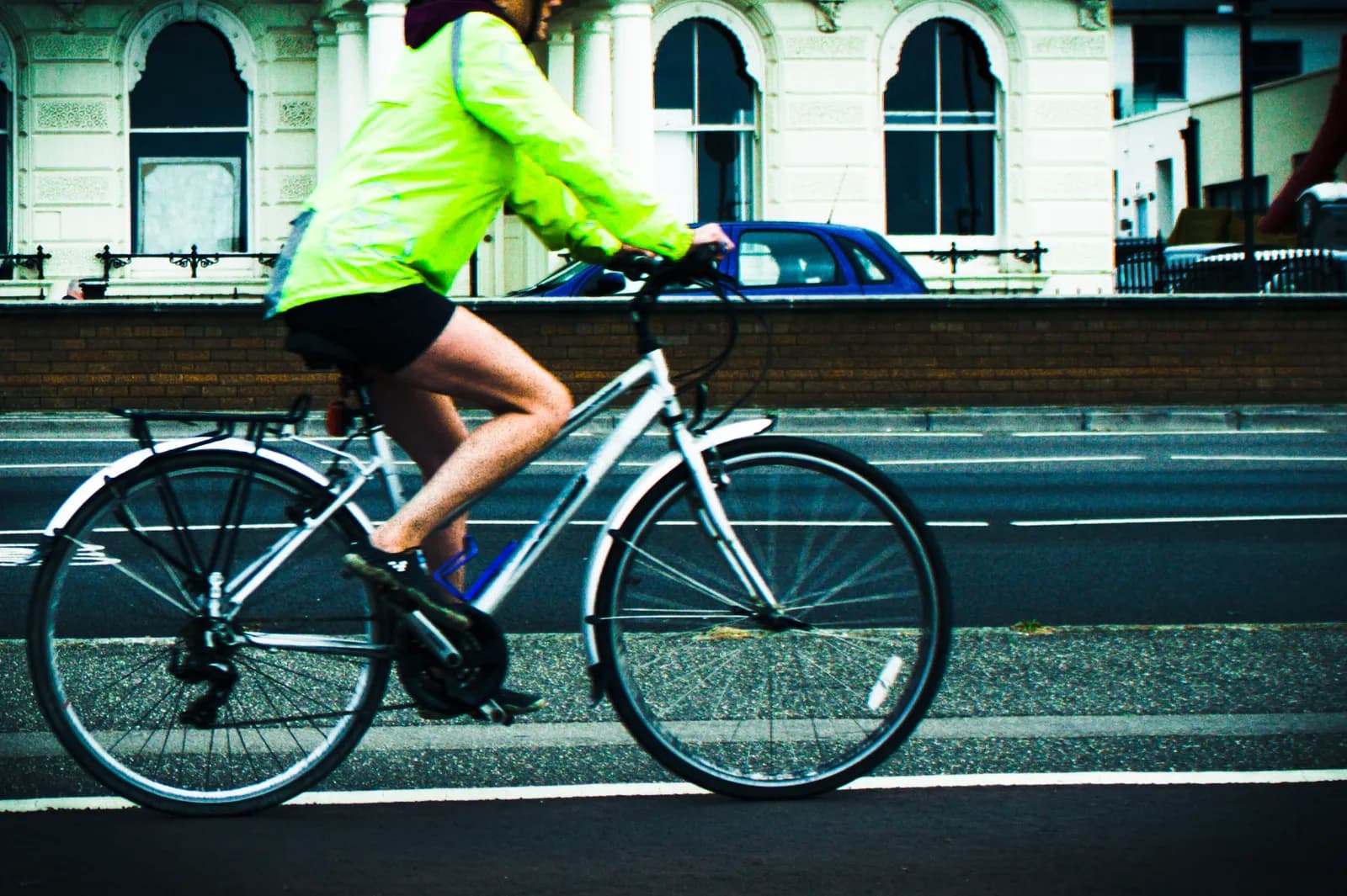 A person in a bright green jacket and black shorts rides a white bicycle on a road with a brick wall and white buildings in the background.