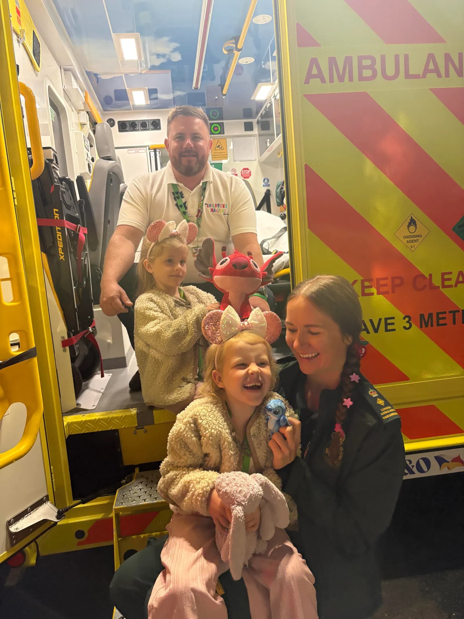 A man in a white polo shirt, two young girls wearing Minnie Mouse headbands, and a woman in a dark uniform with pink star pins in her hair pose by an ambulance.