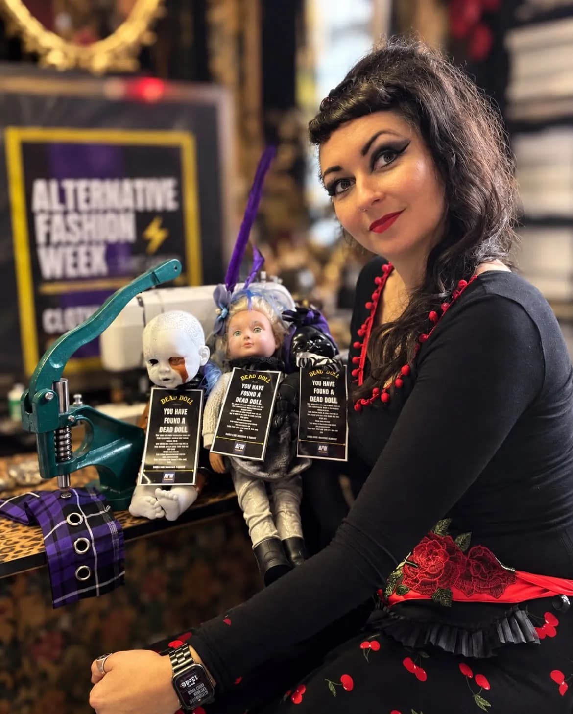 A woman with dark hair and striking makeup smiles, seated next to several creepy dolls with 'DEAD DOLL' tags, with an 'ALTERNATIVE FASHION WEEK' sign in the background.