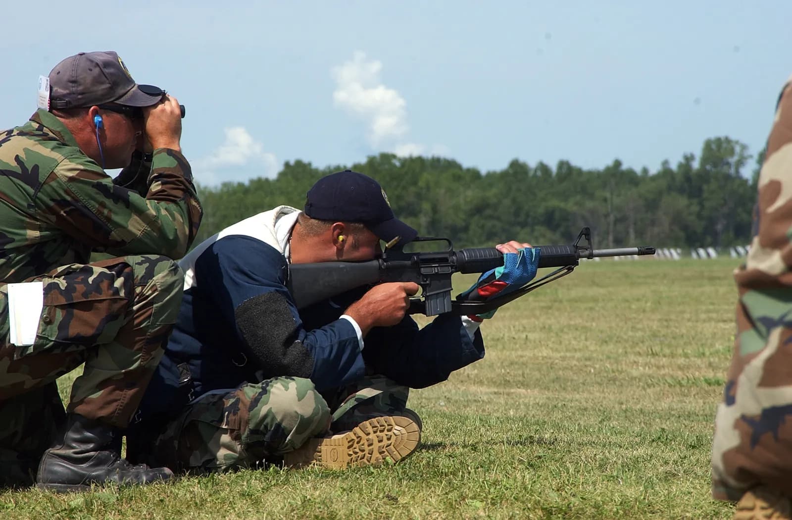 A man in a dark blue shirt and camouflage pants kneels on a grassy field, aiming a rifle, while another man in camouflage kneels behind him, looking through binoculars.