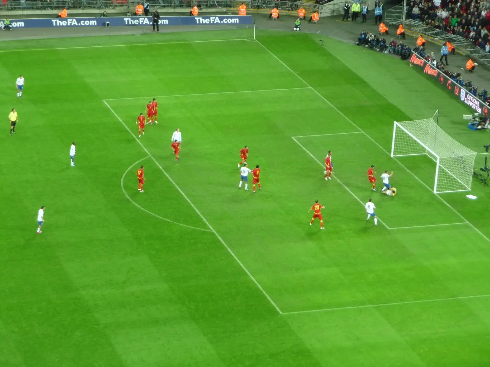 A live soccer game on a green field with players in red and white jerseys, a goal, and spectators in the background.