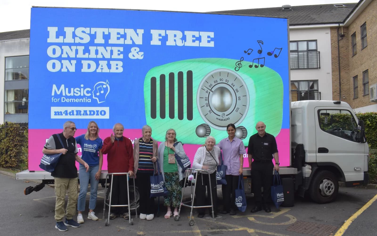 A group of six elderly people, two younger women, and one man stand in front of a truck with a large digital billboard promoting 'Music for Dementia' radio.