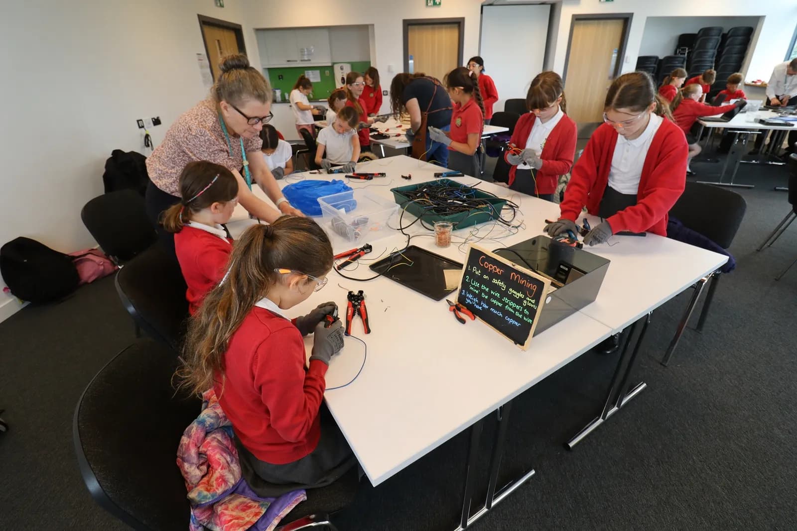 A group of elementary school-aged children and an adult are gathered around tables, engaged in an activity that involves stripping wires, with tools and materials spread across the surfaces.