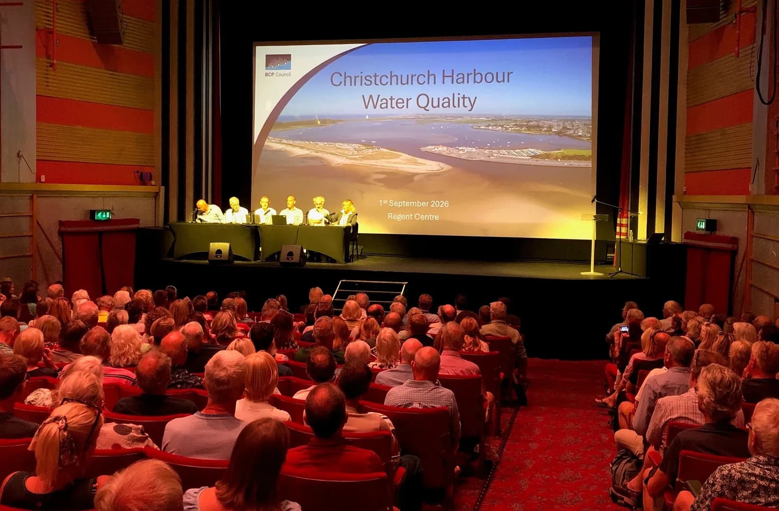 A panel of speakers sitting at a long table on a stage in a large auditorium, presenting to an audience about Christchurch Harbour Water Quality.