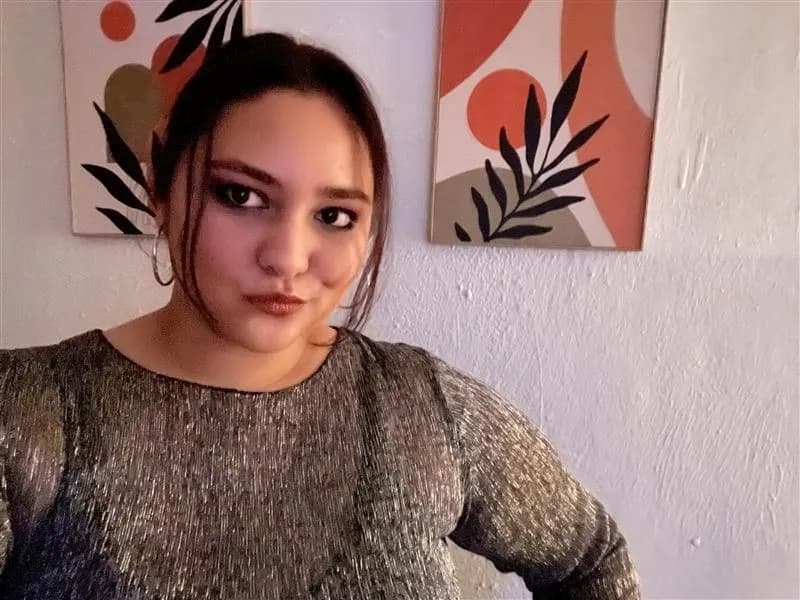 A woman with dark hair and smoky eye makeup wearing a glittering silver top looks directly at the camera with a slight pout, with abstract wall art in the background.