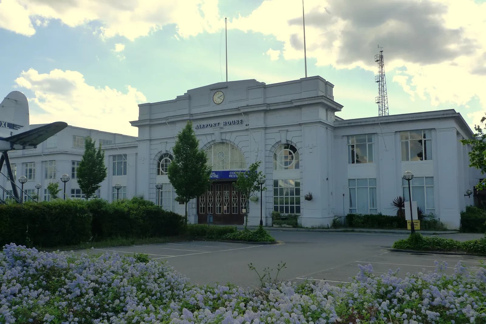 The historic Airport House building, a white art deco structure with a clock tower and arched windows, stands under a cloudy sky, with a low hedge and a parking area in the foreground, and a portion of a plane wing visible on the far left.