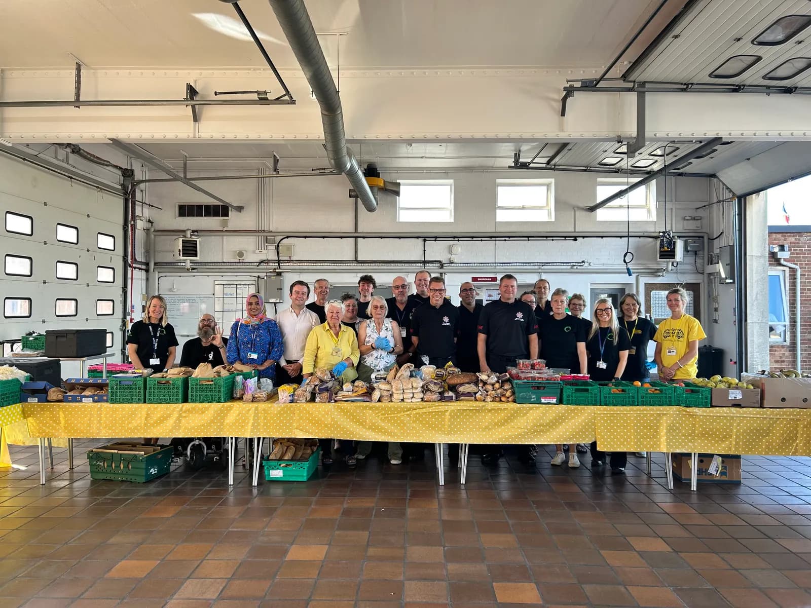 A diverse group of men and women stand behind a long table filled with fresh produce, bread, and groceries, likely at a food bank or community event.