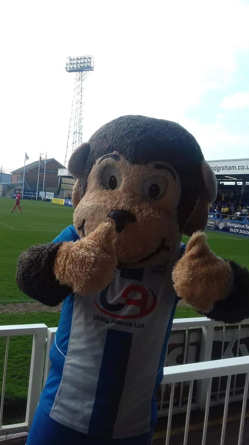 Mascot monkey wearing a sports jersey, making a shushing gesture with one hand over its mouth, standing in front of a sports stadium.