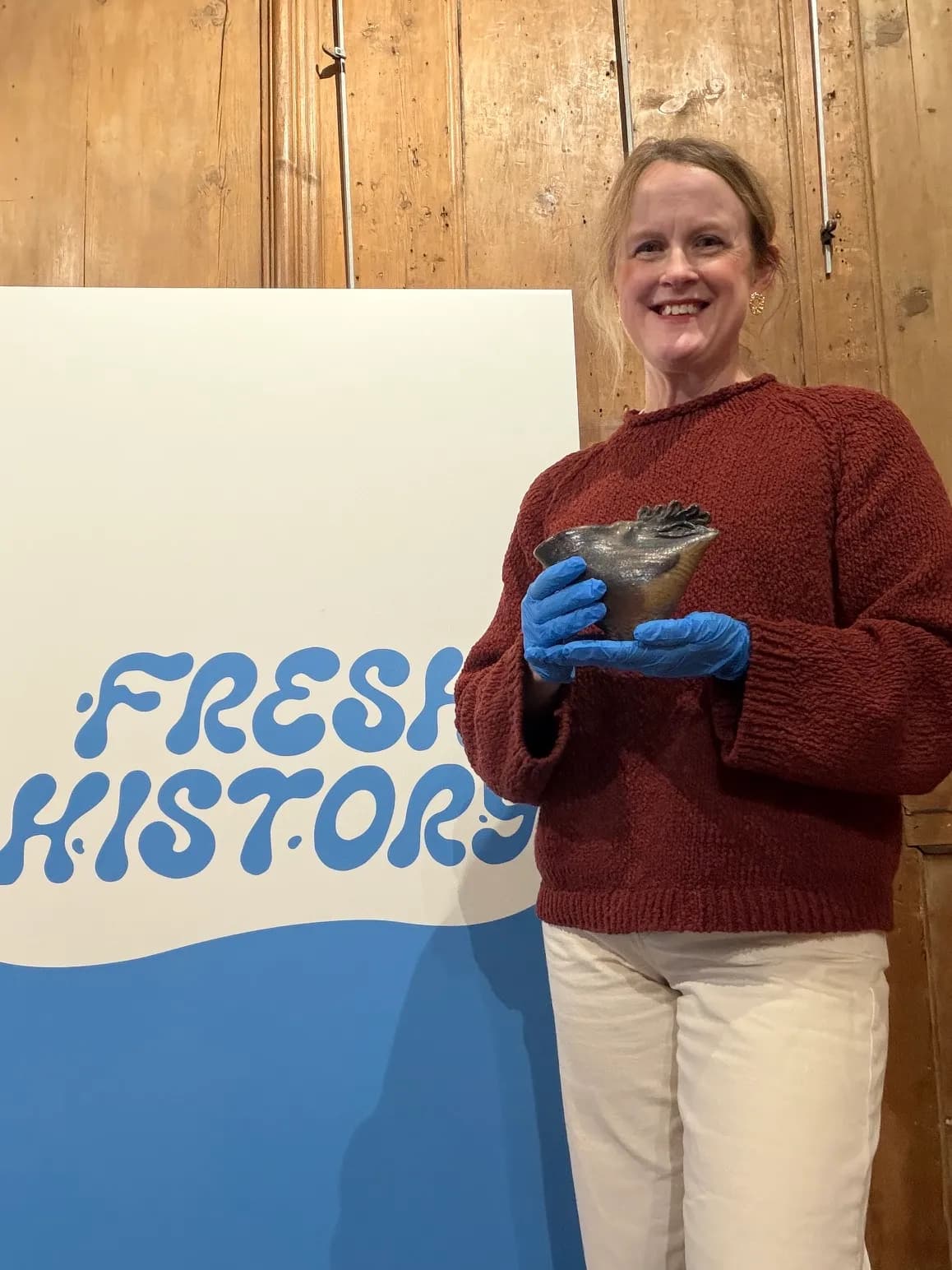 A smiling woman in a maroon sweater and white pants wears blue gloves while holding a small, dark, organic-shaped bowl in front of a blue and white sign that says "FRESH HISTORY."