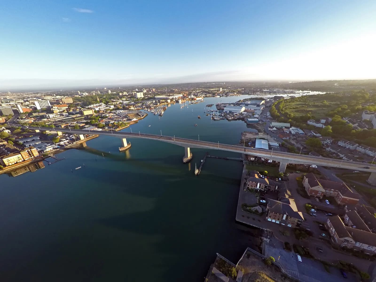 An aerial view of a bridge spanning a wide river with a city and harbor visible on both sides under a clear blue sky.