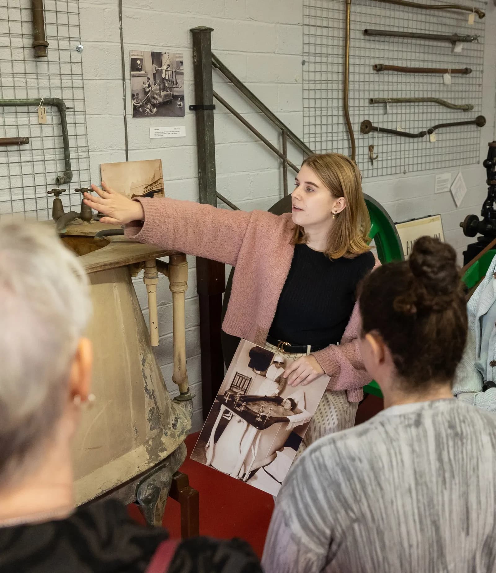 A woman in a pink sweater and black shirt gestures with her right hand towards an old washbasin while holding a historical photograph, as two other individuals listen.