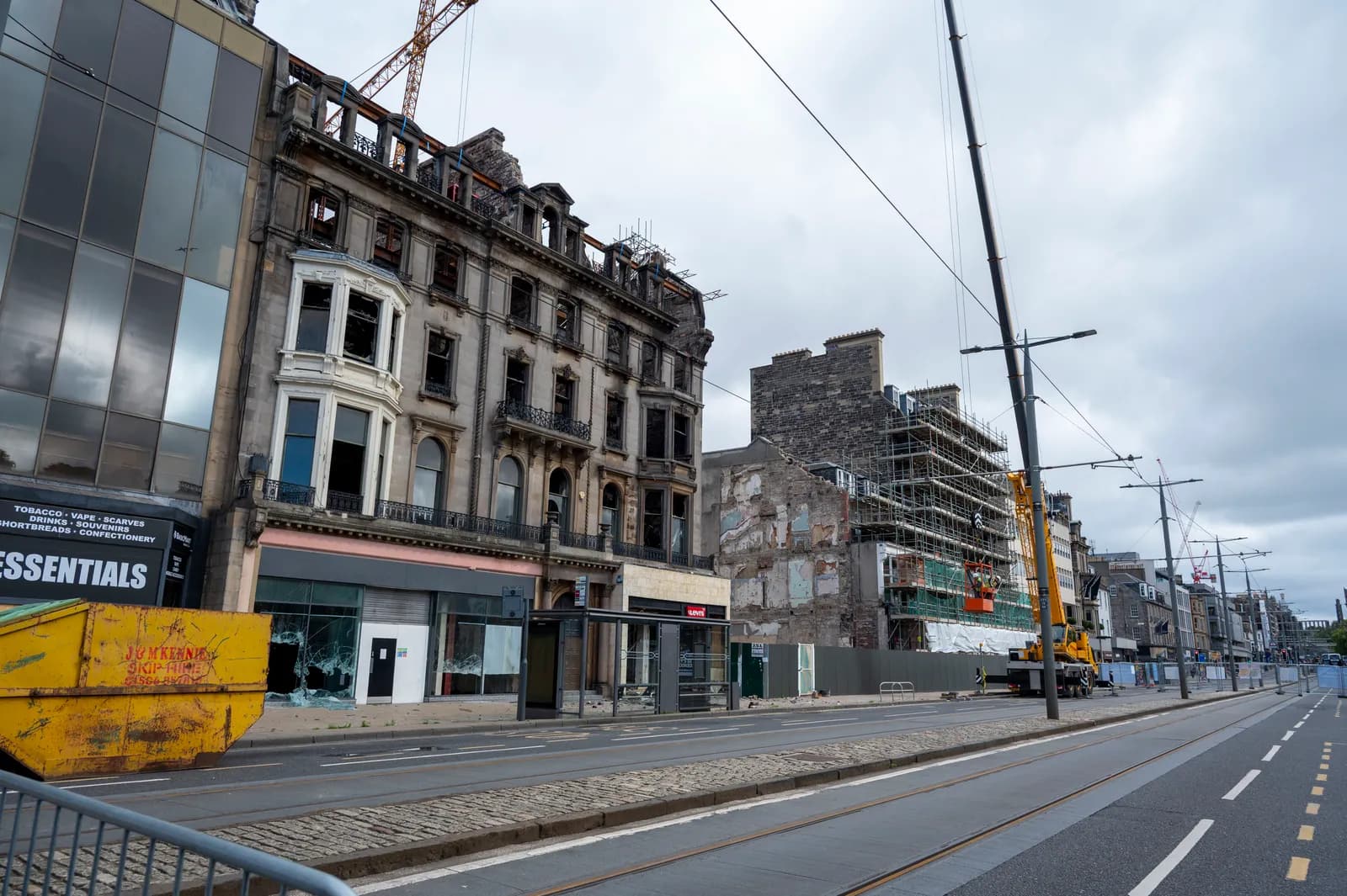 A street scene featuring old, damaged buildings, some under construction with scaffolding, alongside a tram line and a large yellow skip.