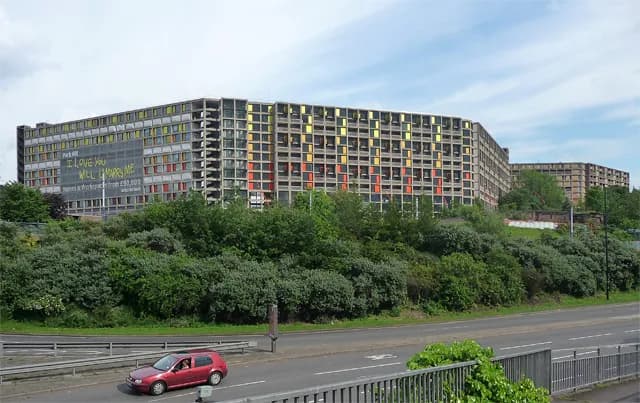 A long, concrete apartment building with many square windows in bright colors on a lush, green hill under a cloudy sky.