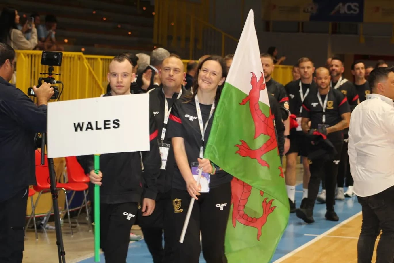 A group of people wearing black activewear, one holding a sign that says "WALES," and another holding a Welsh flag, walk through an arena.