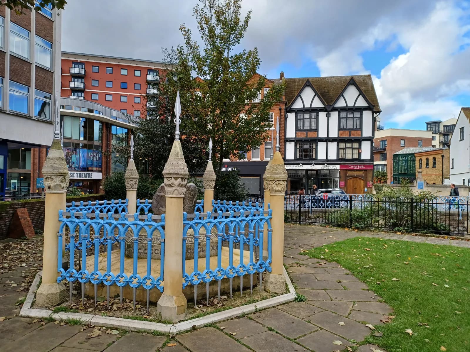 A small blue decorative fence surrounds a stone monument in a paved area, with historical and modern buildings in the background under a cloudy sky.