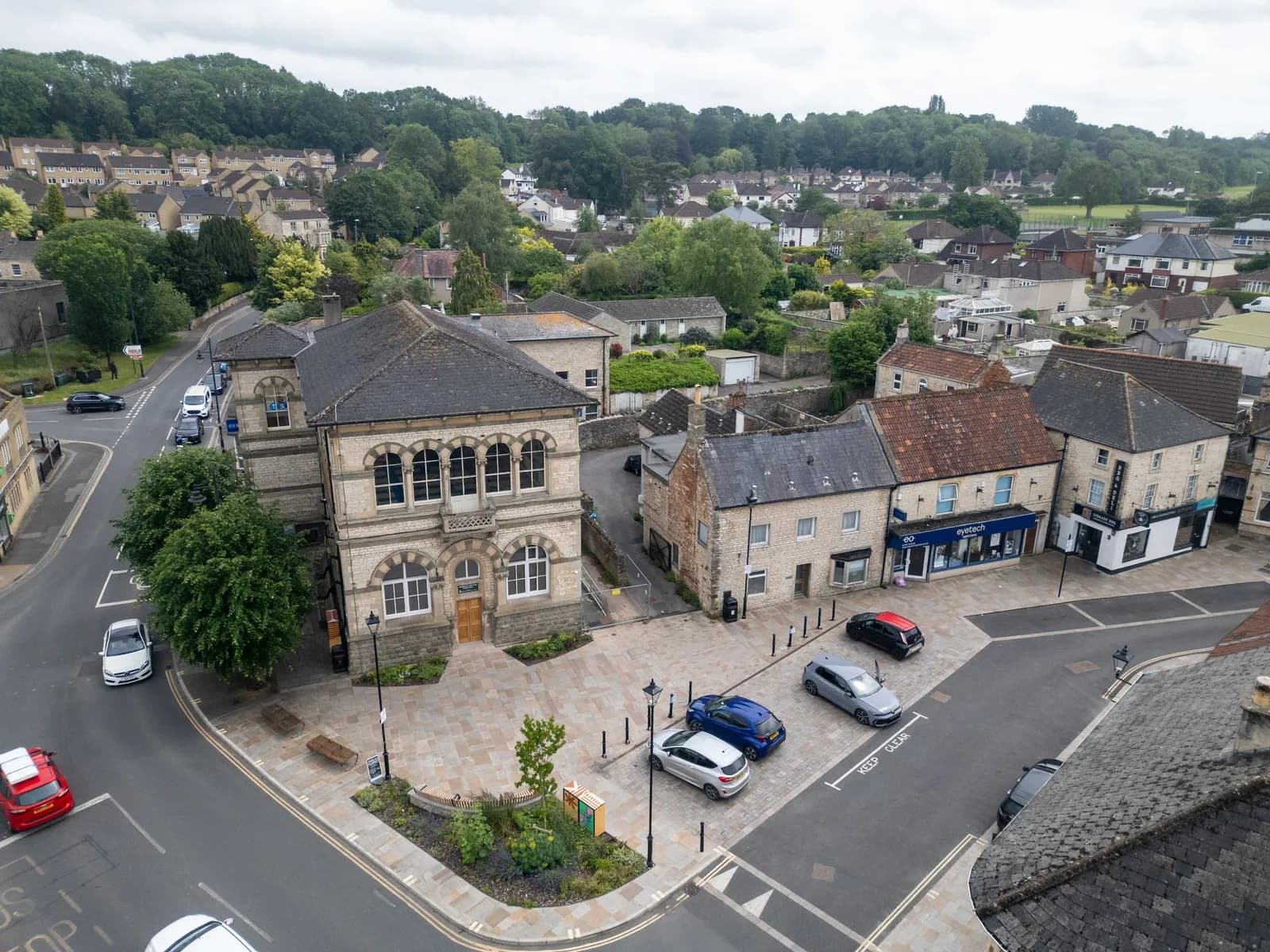 An aerial view of a town square with historic buildings and parked cars, surrounded by residential areas and lush green hills under a cloudy sky.