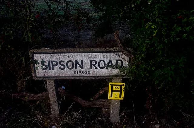 A weather-beaten street sign for Sipson Road, partially obscured by dark foliage, with a yellow hydrant marker below it.