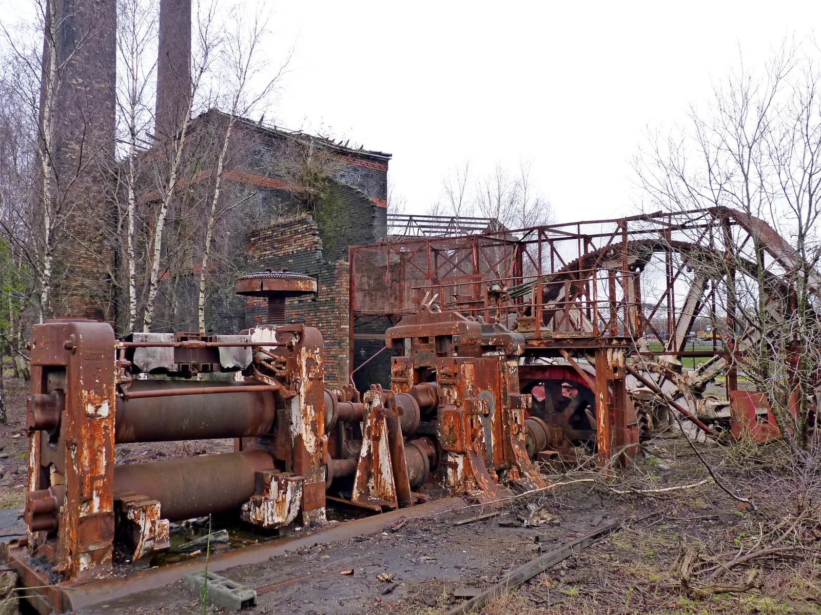 Rusting industrial machinery stands in the foreground, with two tall brick chimney stacks and a dilapidated brick building in the background, surrounded by bare trees under a light sky.