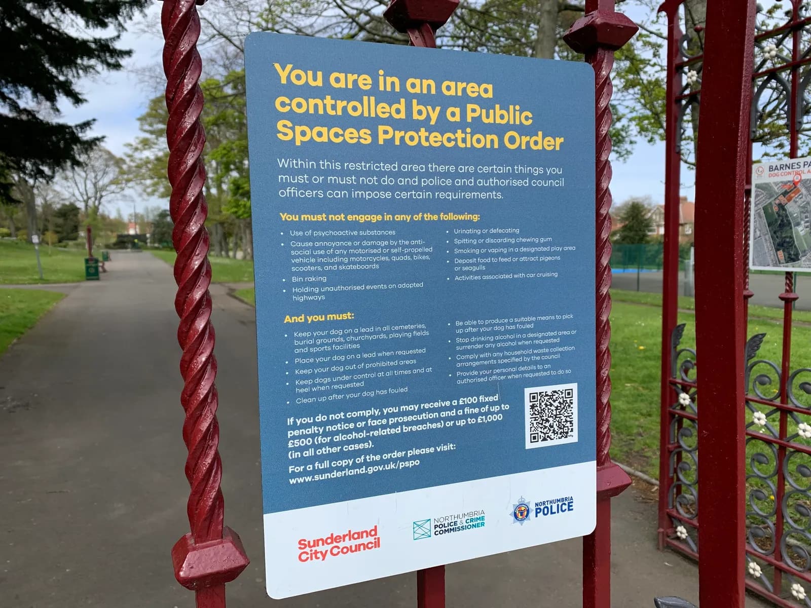 A Public Spaces Protection Order sign on a dark red metal gate, detailing regulations for a public area.