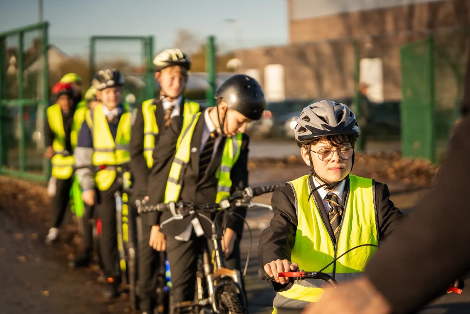 A group of school children in helmets and high-visibility vests push their bikes outdoors, with the child in the foreground looking towards the right.