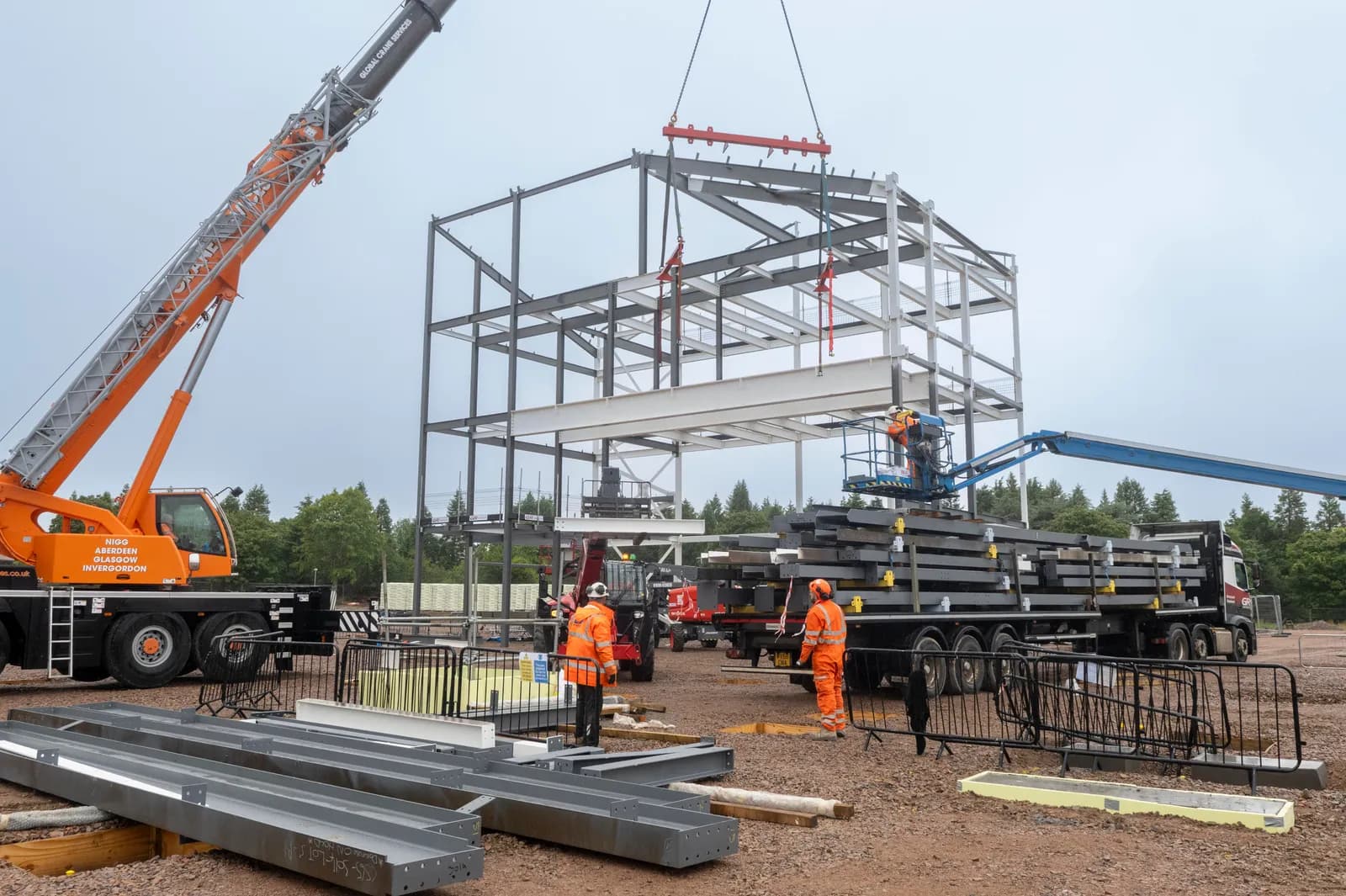 A large construction crane lifting a white steel beam into place on a multi-story metal framework, with several construction workers in orange high-visibility clothing nearby.