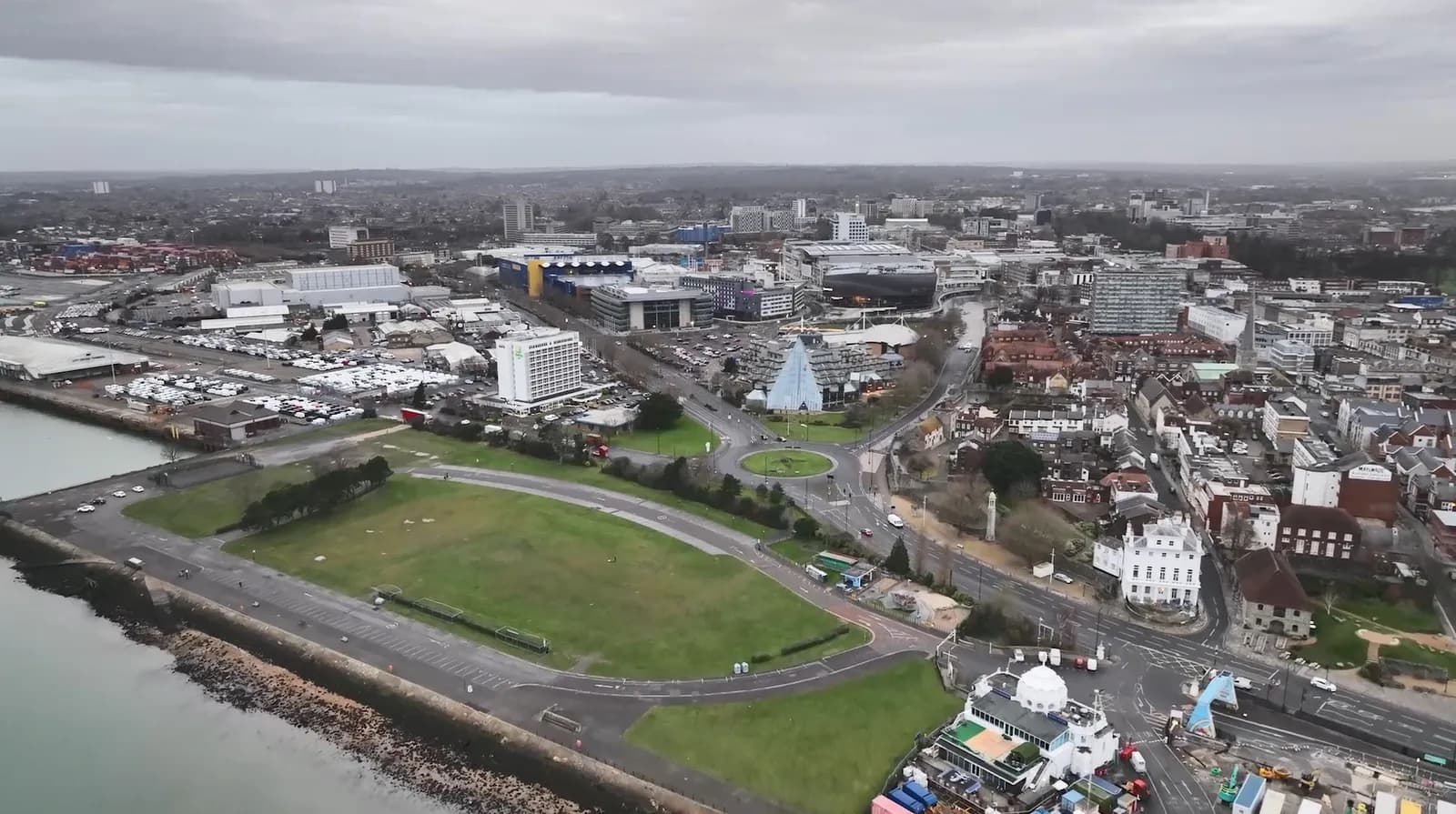 An aerial view of a coastal city under an overcast sky, showing a mix of commercial buildings, residential areas, green spaces, and a large roundabout.
