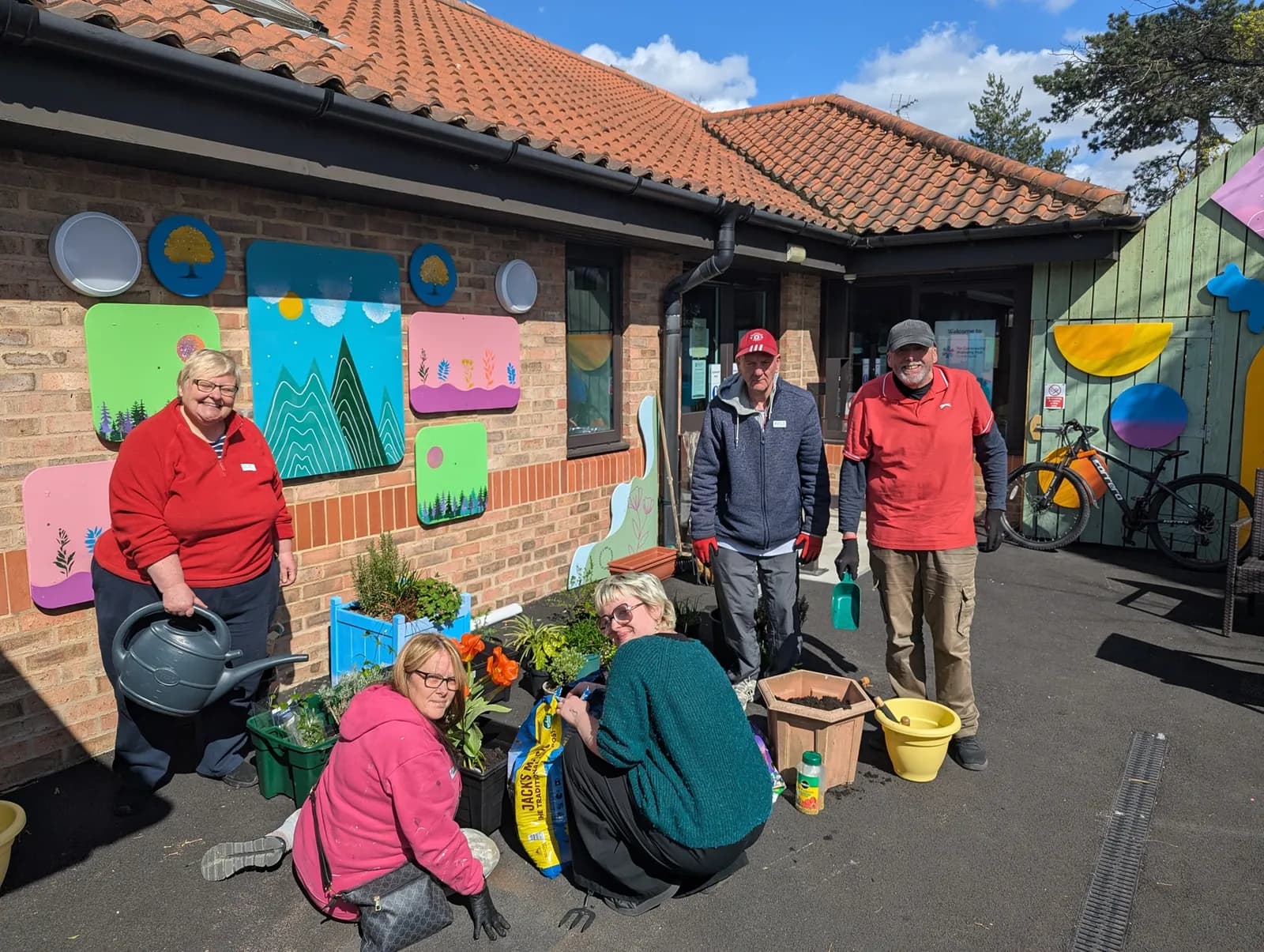 A group of five people are gardening outdoors near a brick building with colorful decorative panels on its wall.