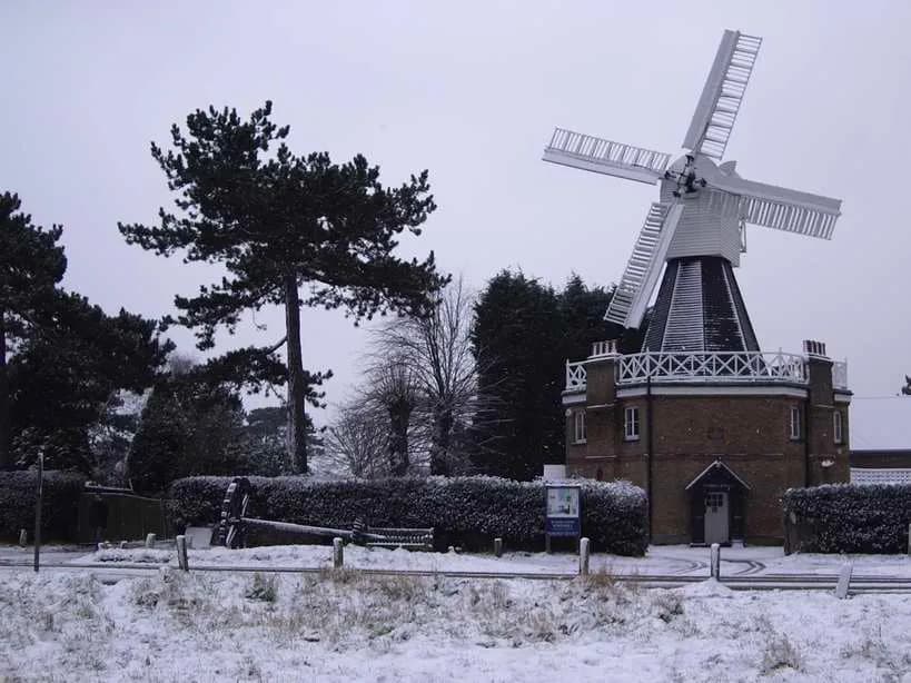 A historic brick windmill with white sails stands prominent against a grey, snowy sky, surrounded by snow-dusted trees and ground.