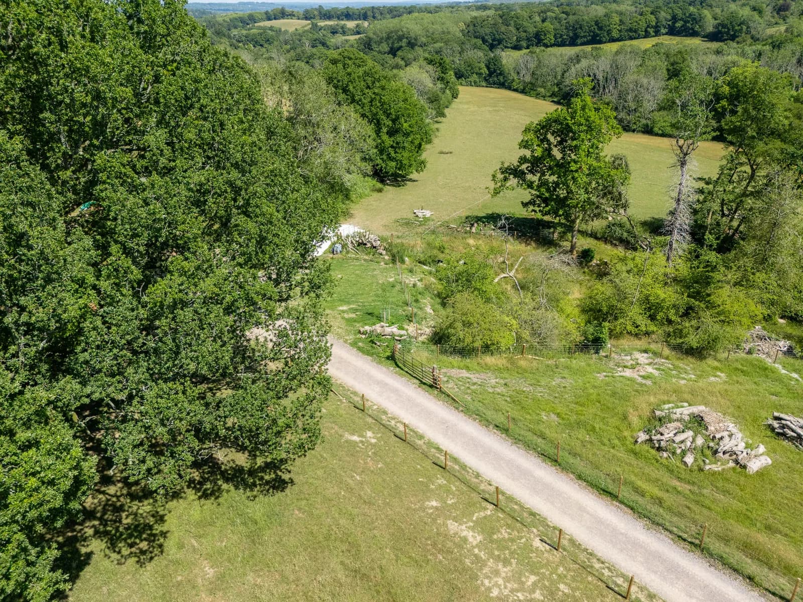 An aerial view of a winding dirt road leading through green fields and dense forests under a bright sky.