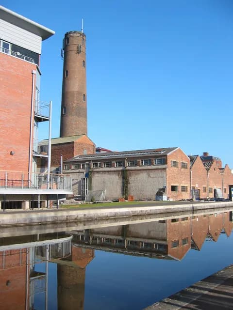 A tall brick tower stands behind a red-brick building, bordering a canal whose still water reflects the structures and clear blue sky.
