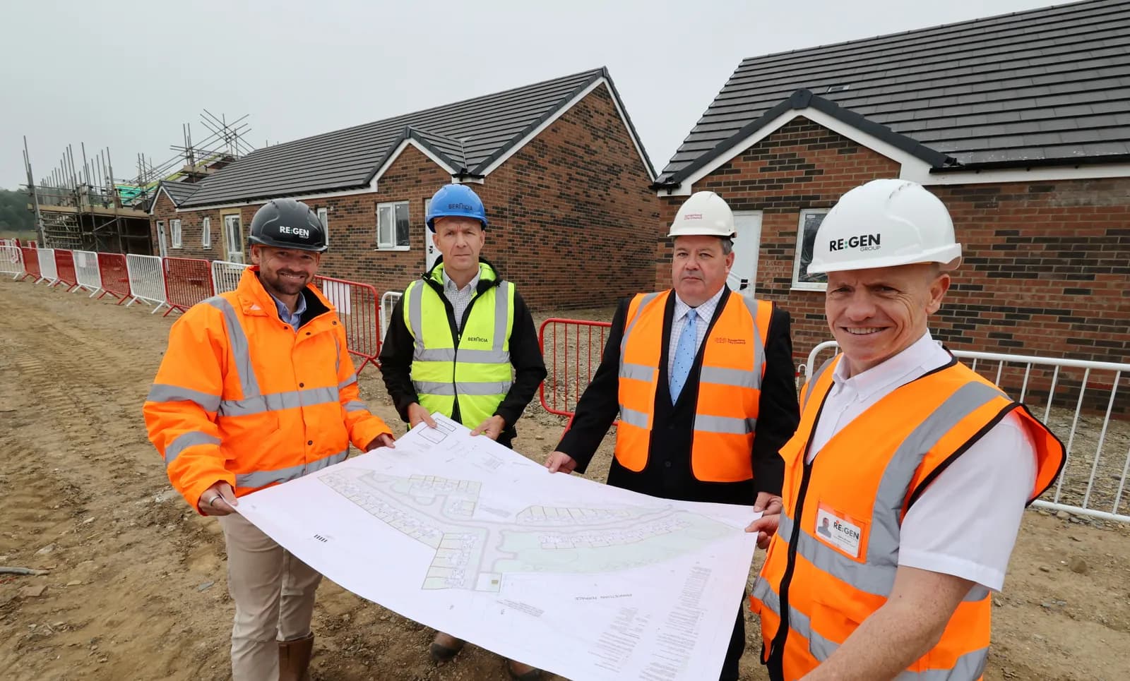 Four men in hard hats and high-visibility vests examine a large blueprint at a construction site with new brick houses in the background.