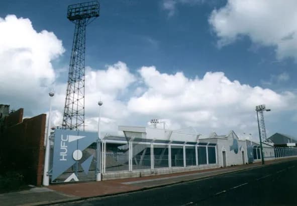 A tall stadium light structure looms over a building with a blue 'HUFC' sign and a long street running alongside it under a cloudy sky.