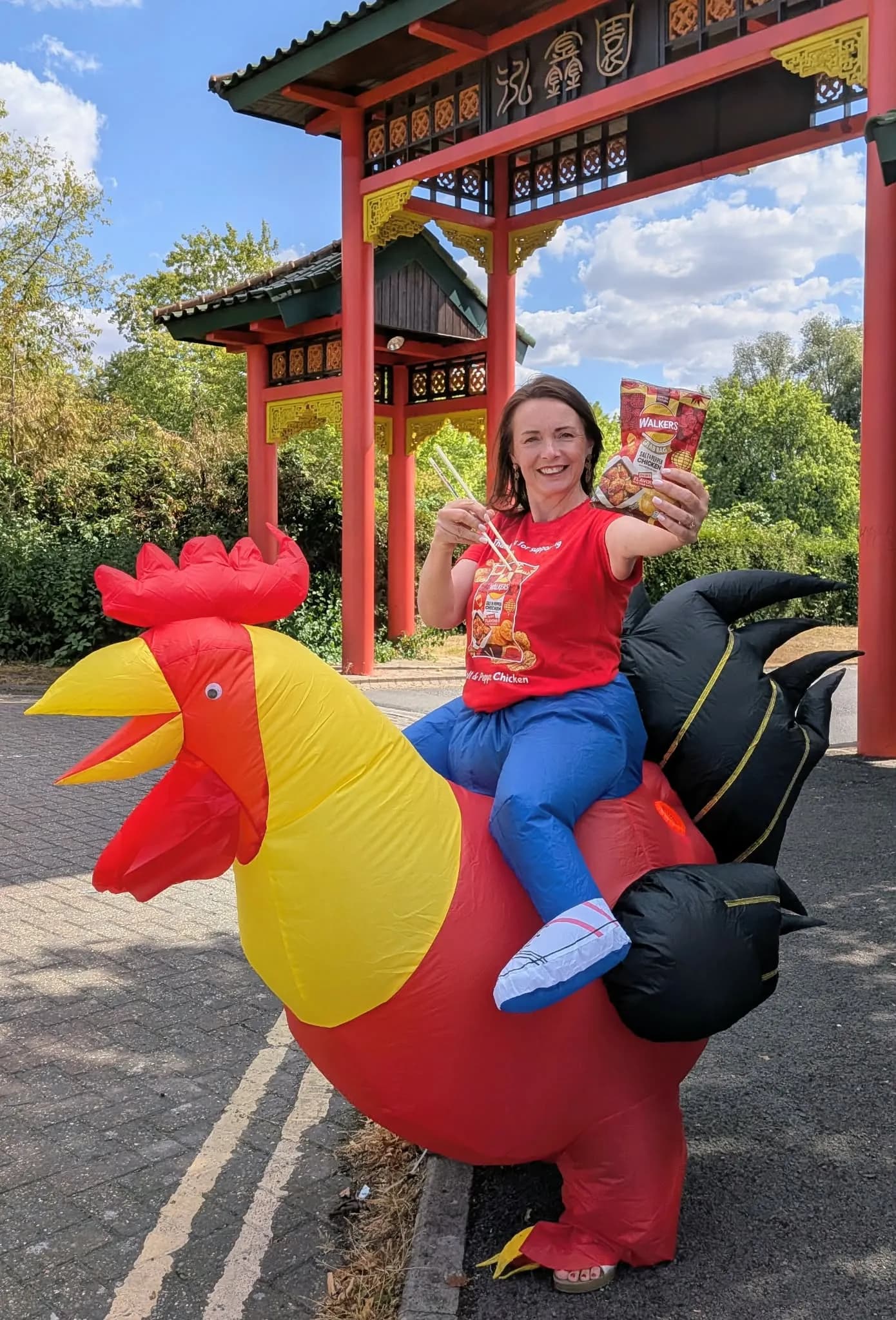 A woman dressed in a chicken rider inflatable costume holds up a packet of Walkers crisps and chopsticks, smiling in front of a red Chinese archway.