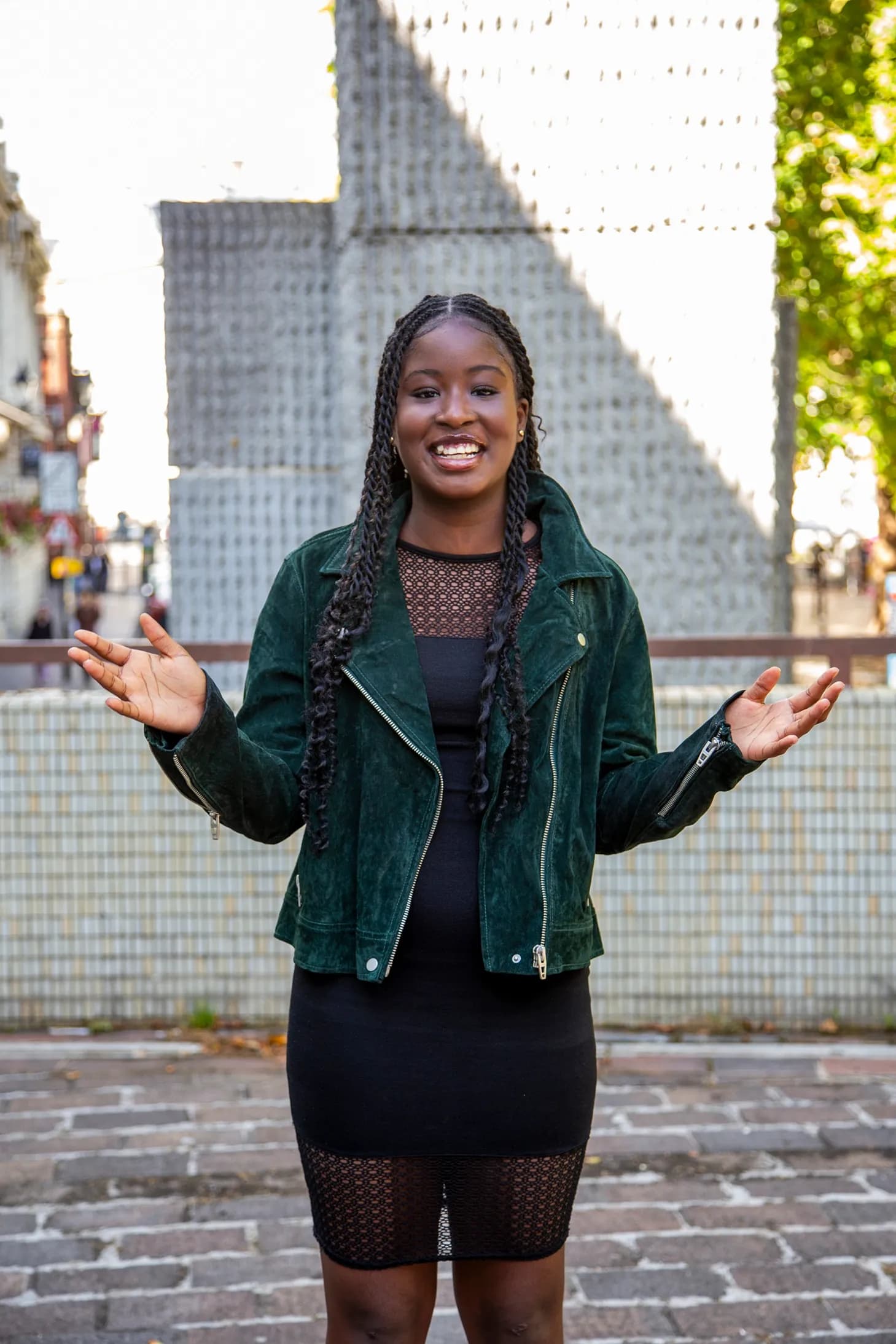 A young woman with braids and a dark green jacket smiles at the camera with her hands outstretched, against a backdrop of a stone wall and blurred street.