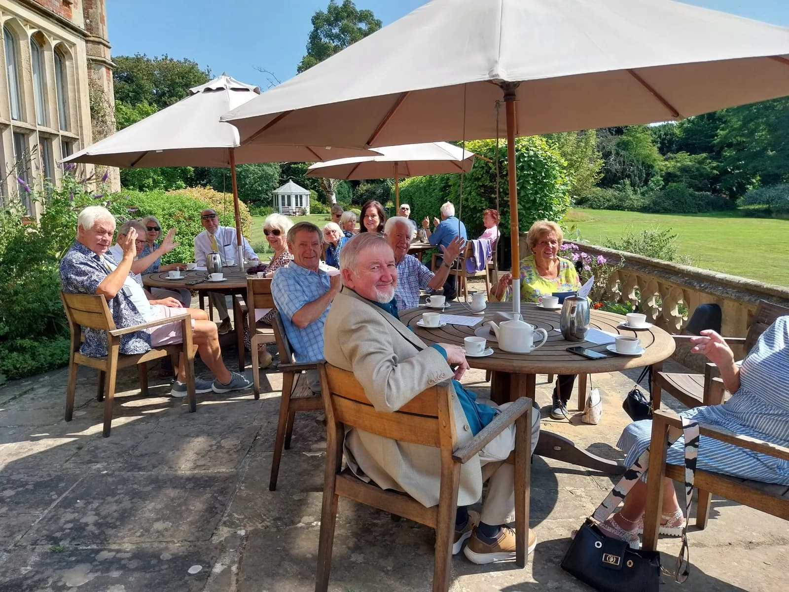 A group of older adults enjoy tea and conversation on a patio shaded by large umbrellas, with a grand building and green lawn in the background.