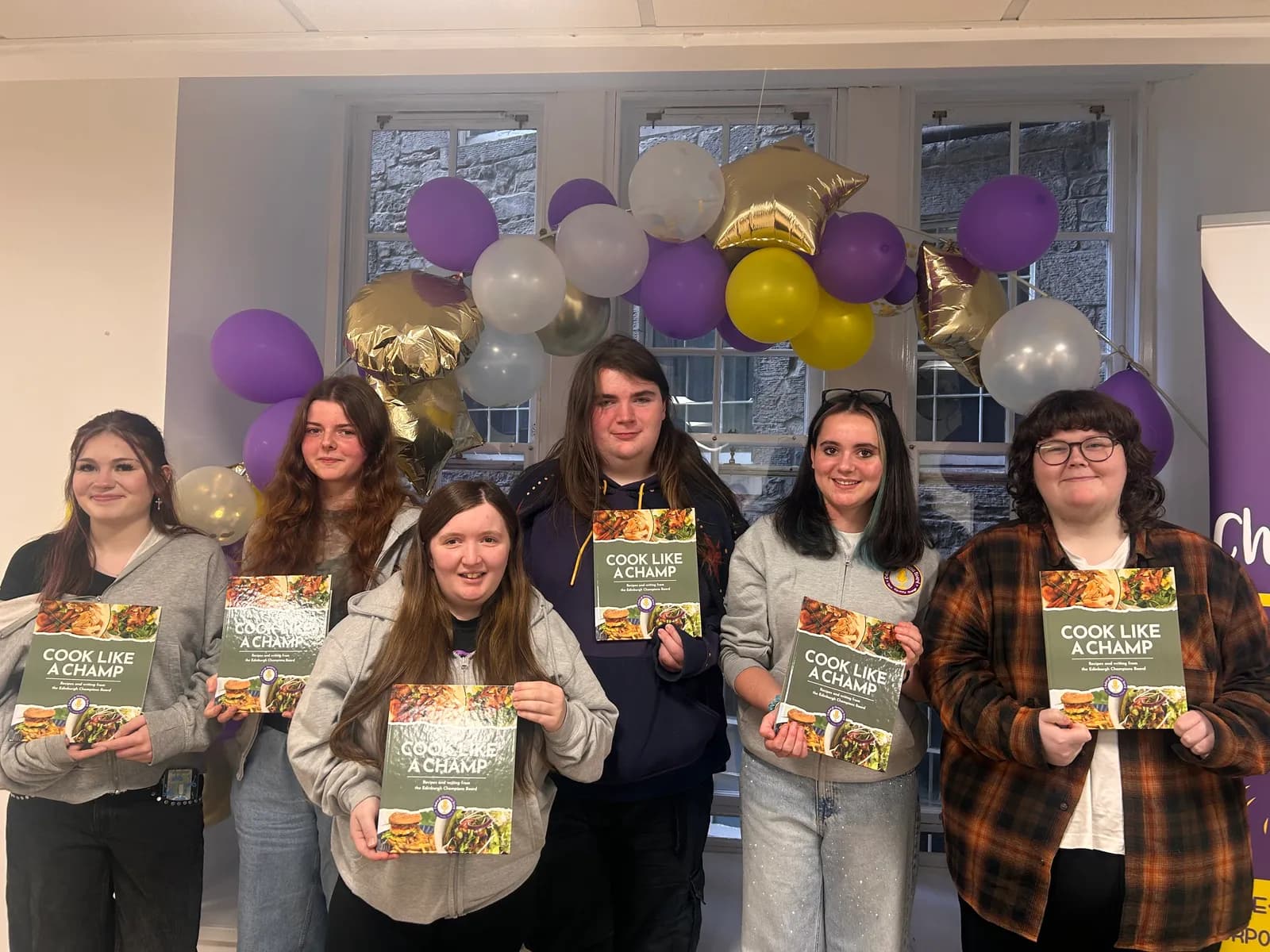 Six young adults pose with copies of a cookbook titled "Cook Like a Champ" in front of a window decorated with purple, yellow, and silver balloons.
