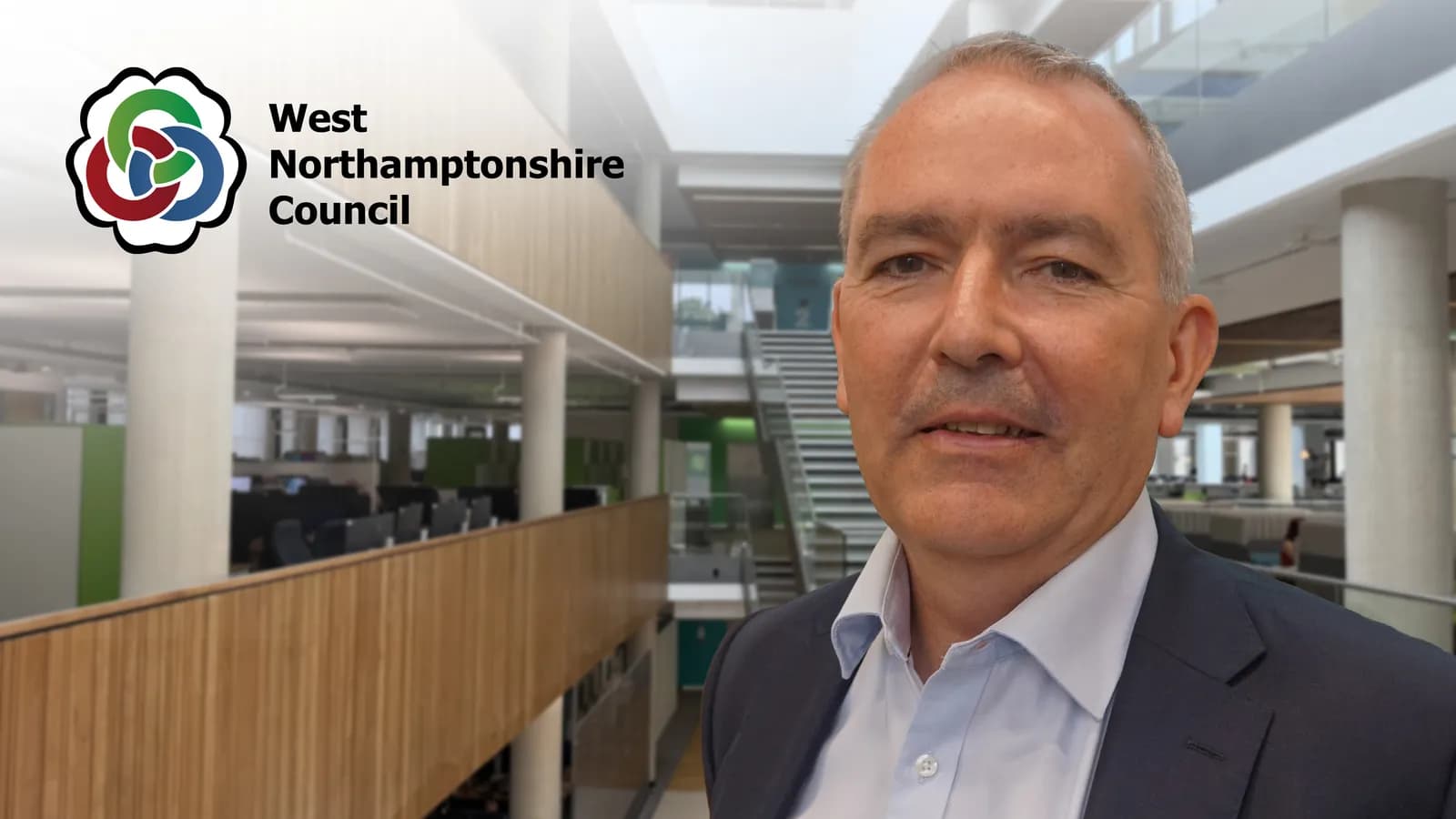 A man in a suit jacket and light blue shirt smiles looking slightly to his left, with a modern office building interior and the West Northamptonshire Council logo in the background.