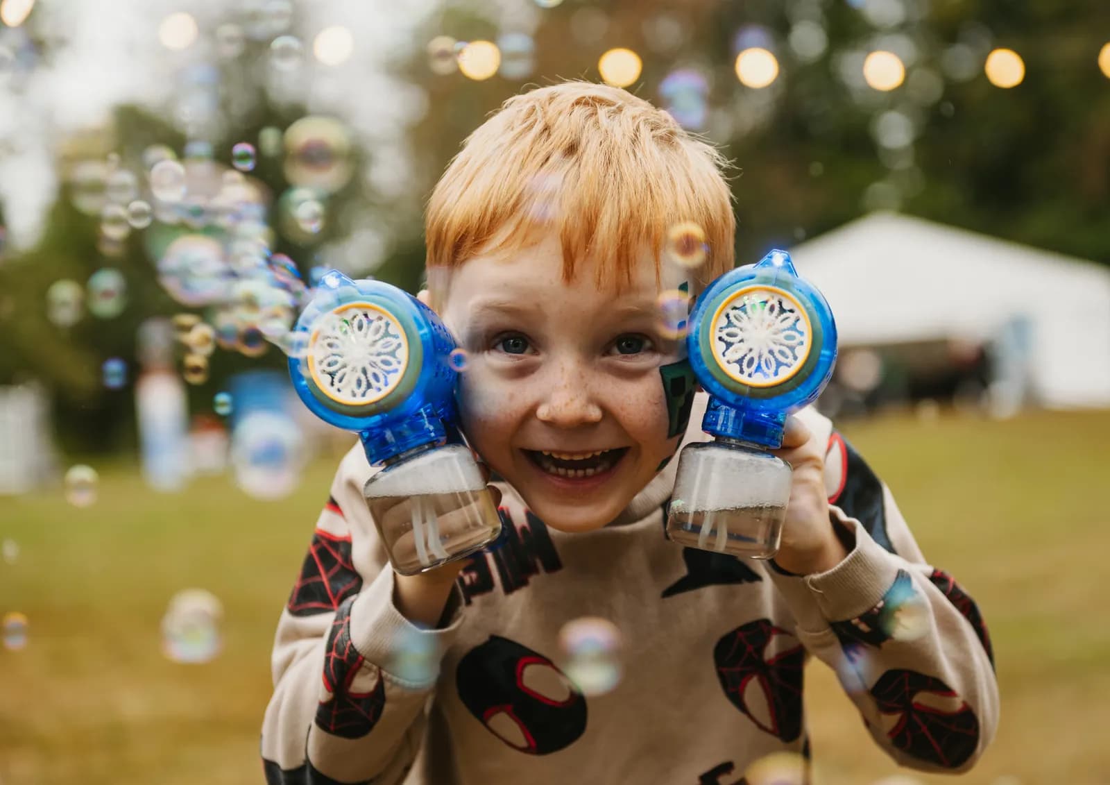 A young boy with red hair smiles broadly while holding two blue bubble guns that are actively producing bubbles, with bokeh lights in the background.