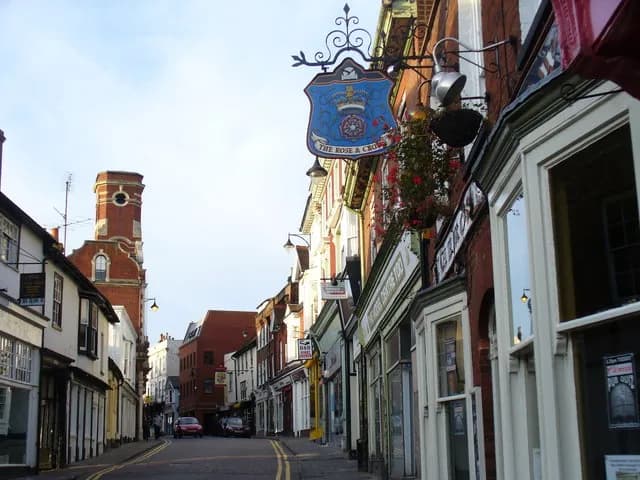 A narrow street lined with buildings, featuring a blue and gold pub sign for "The Rose & Crown" hanging prominently on the right, and a brick tower in the distance.