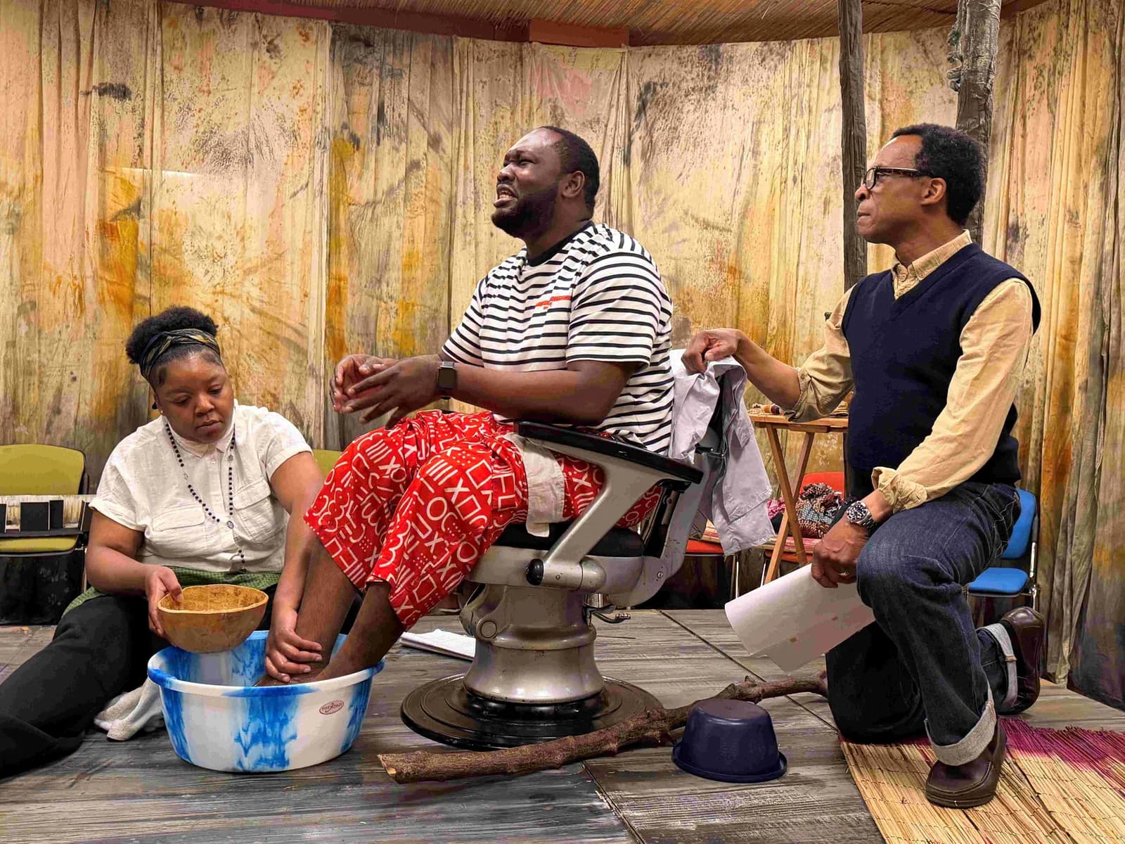 Three people in a theatrical setting; a woman on the left washing a man's feet that are in a basin, while another man on the right holds papers and watches them.