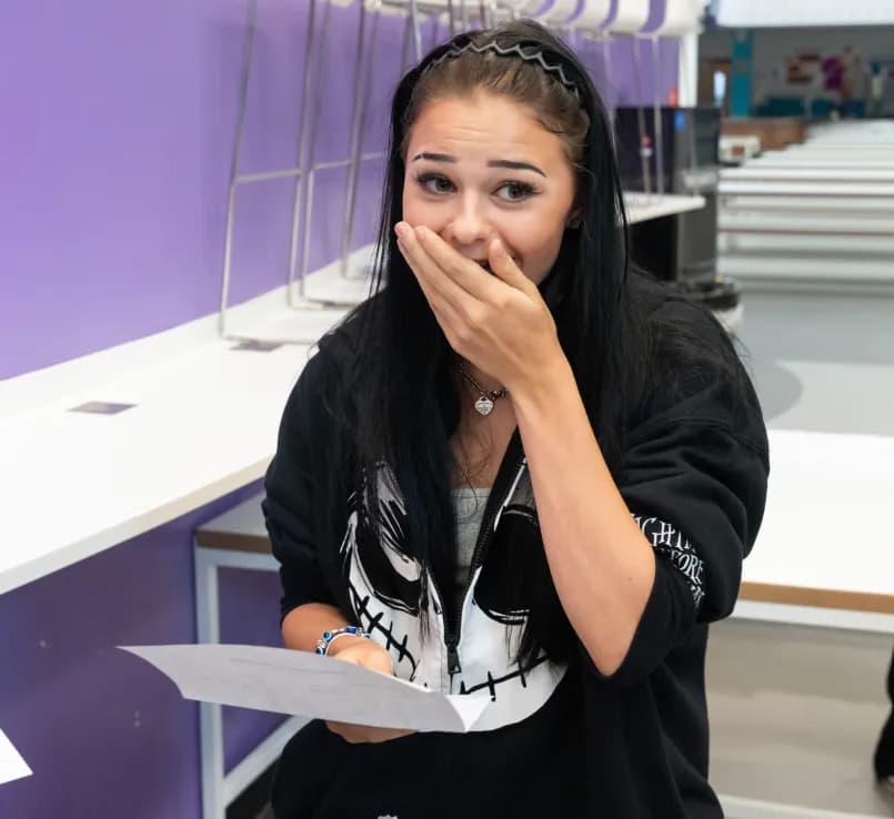 A young woman with long dark hair, wearing a black hoodie, looks surprised with her hand covering her mouth while holding a paper.