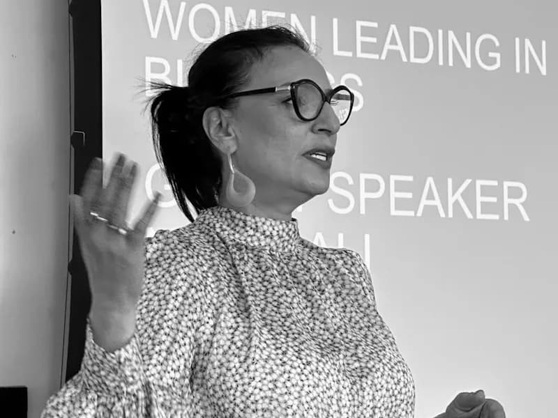 A woman with glasses and a patterned blouse speaks, gesturing with one hand, in front of a screen displaying text that includes "WOMEN LEADING IN" and "SPEAKER."