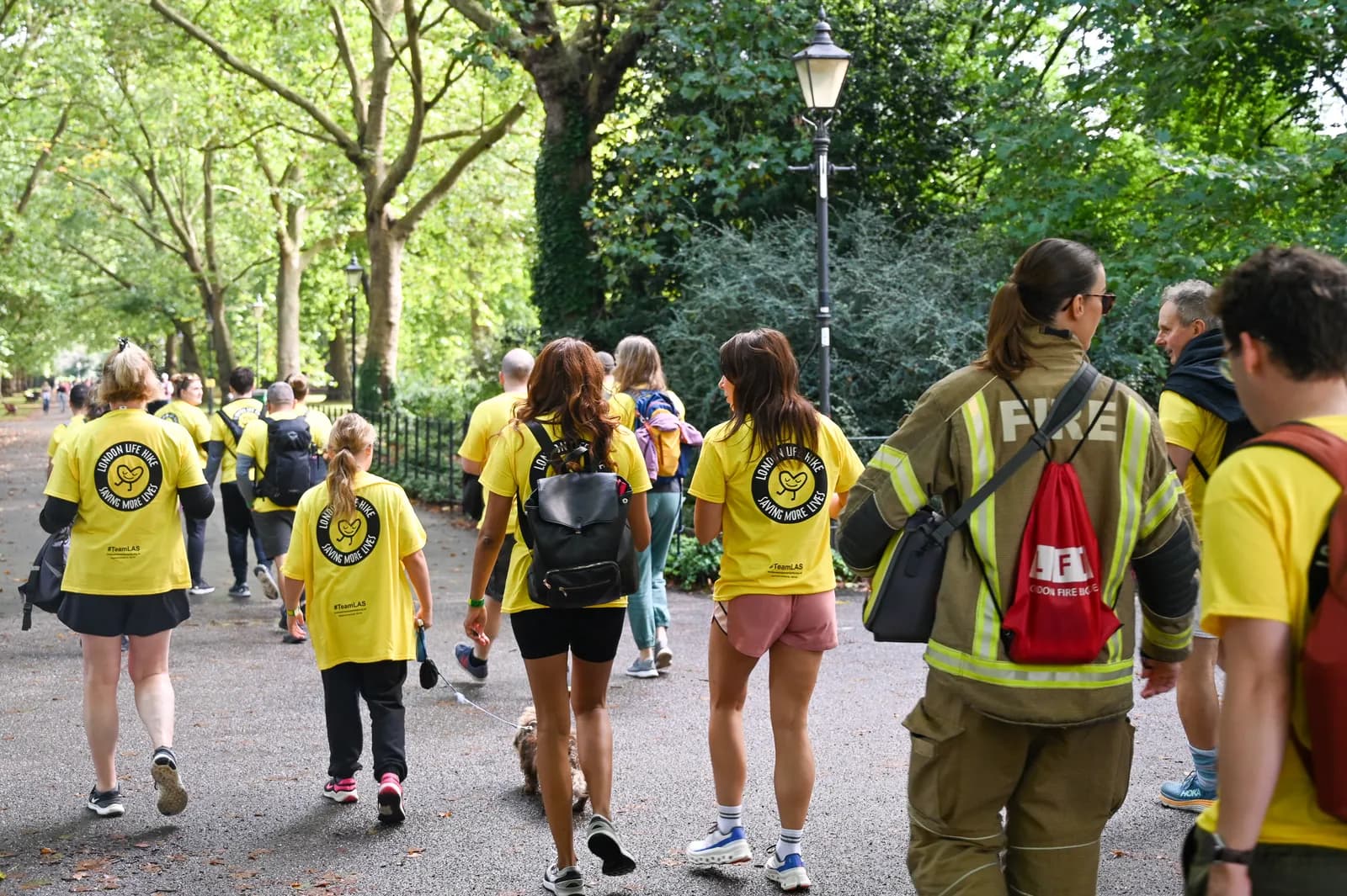 A group of people, many wearing yellow shirts with a heart logo and the words "London Life Hike," walk through a park.