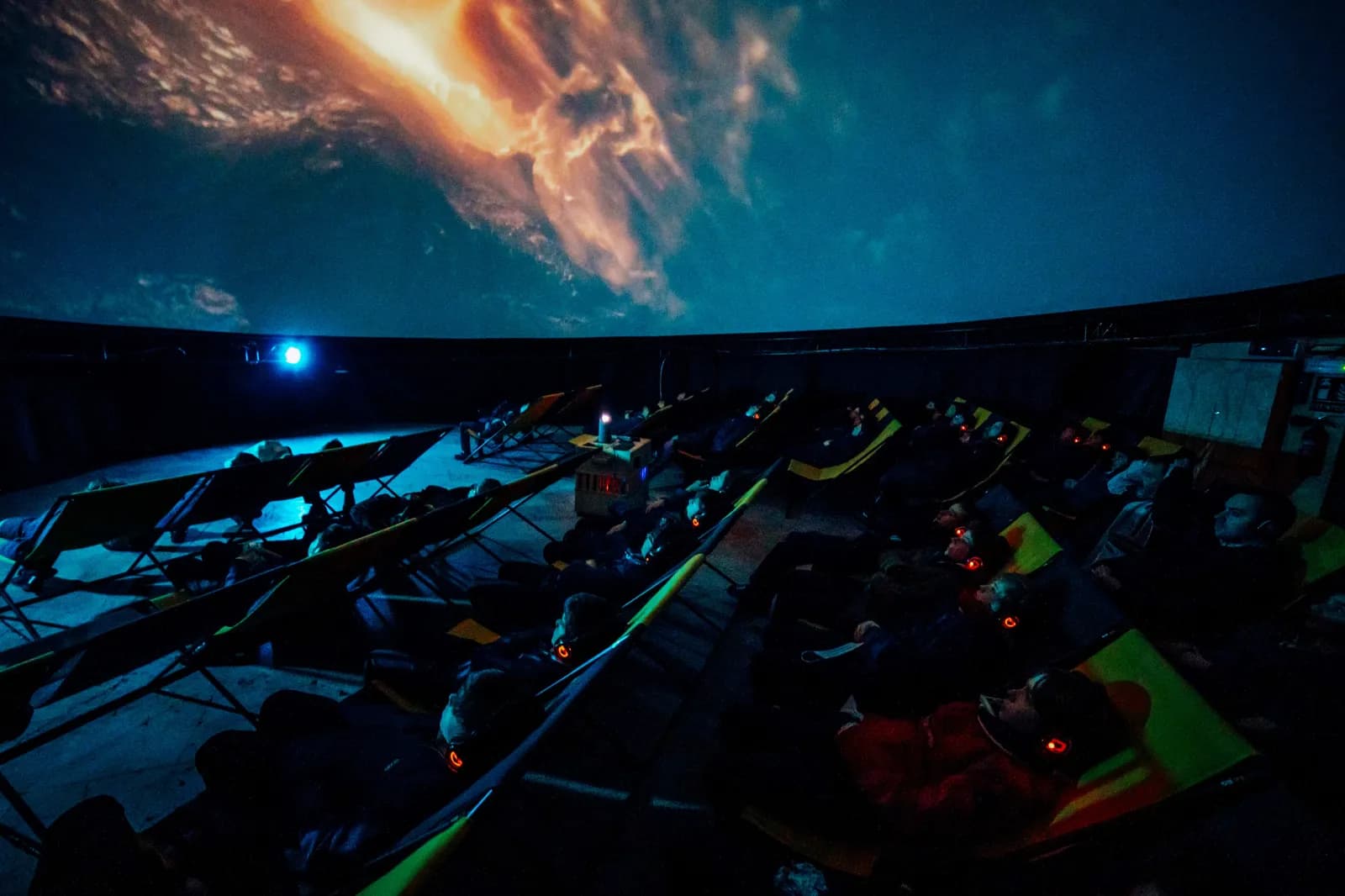 People reclining in chairs under a planetarium dome displaying a cosmic scene with bright orange and blue hues.