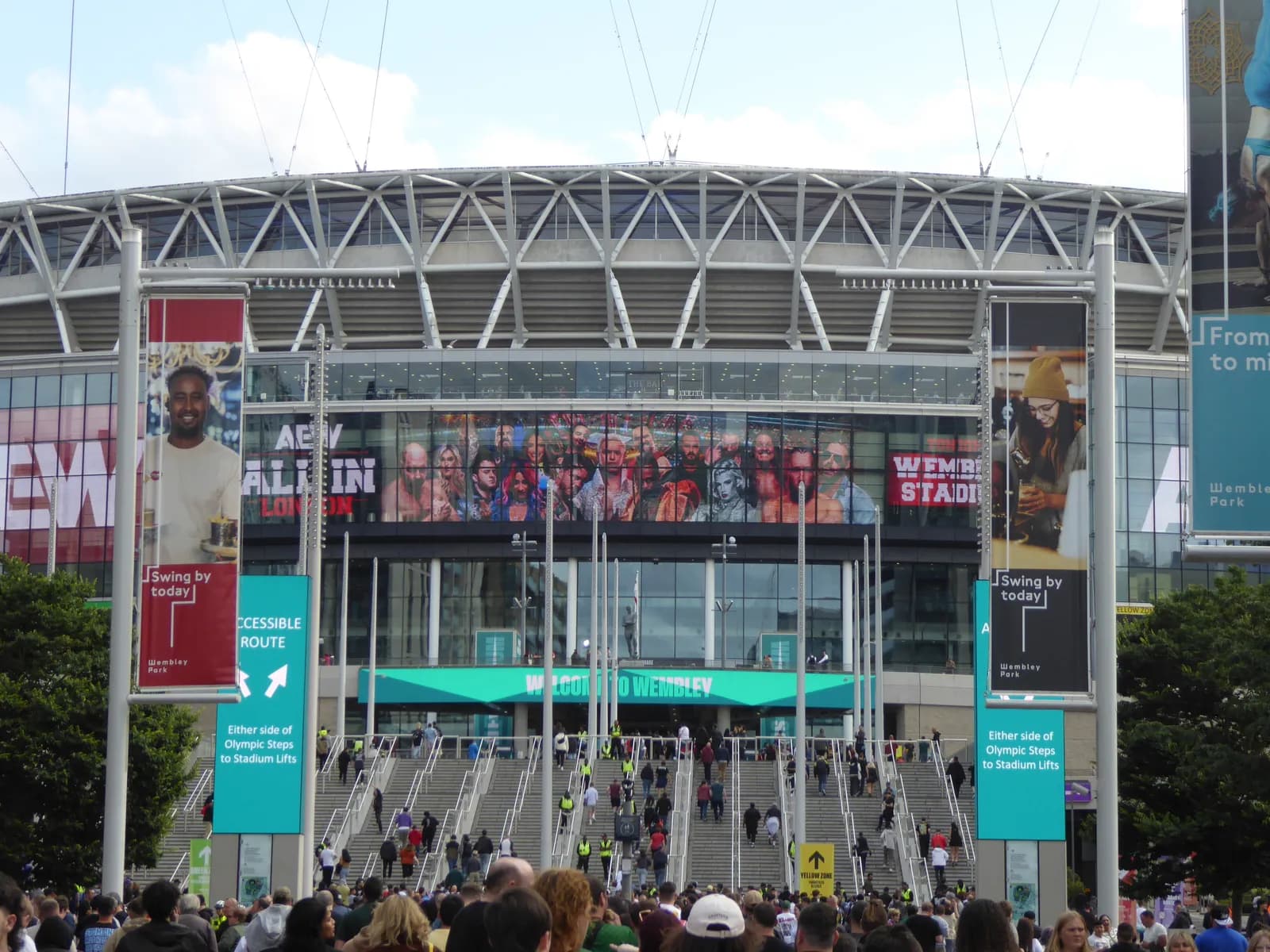 The entrance to Wembley Stadium, with crowds of people walking up concrete stairs and multiple large advertising screens visible on the building.