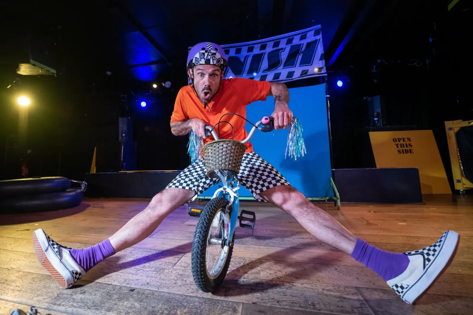 A man wearing an orange shirt, checkerboard shorts, a purple helmet, and checkerboard shoes sits on a small blue bicycle with his legs splayed wide, looking at the camera with a surprised expression on a stage with a blue background.